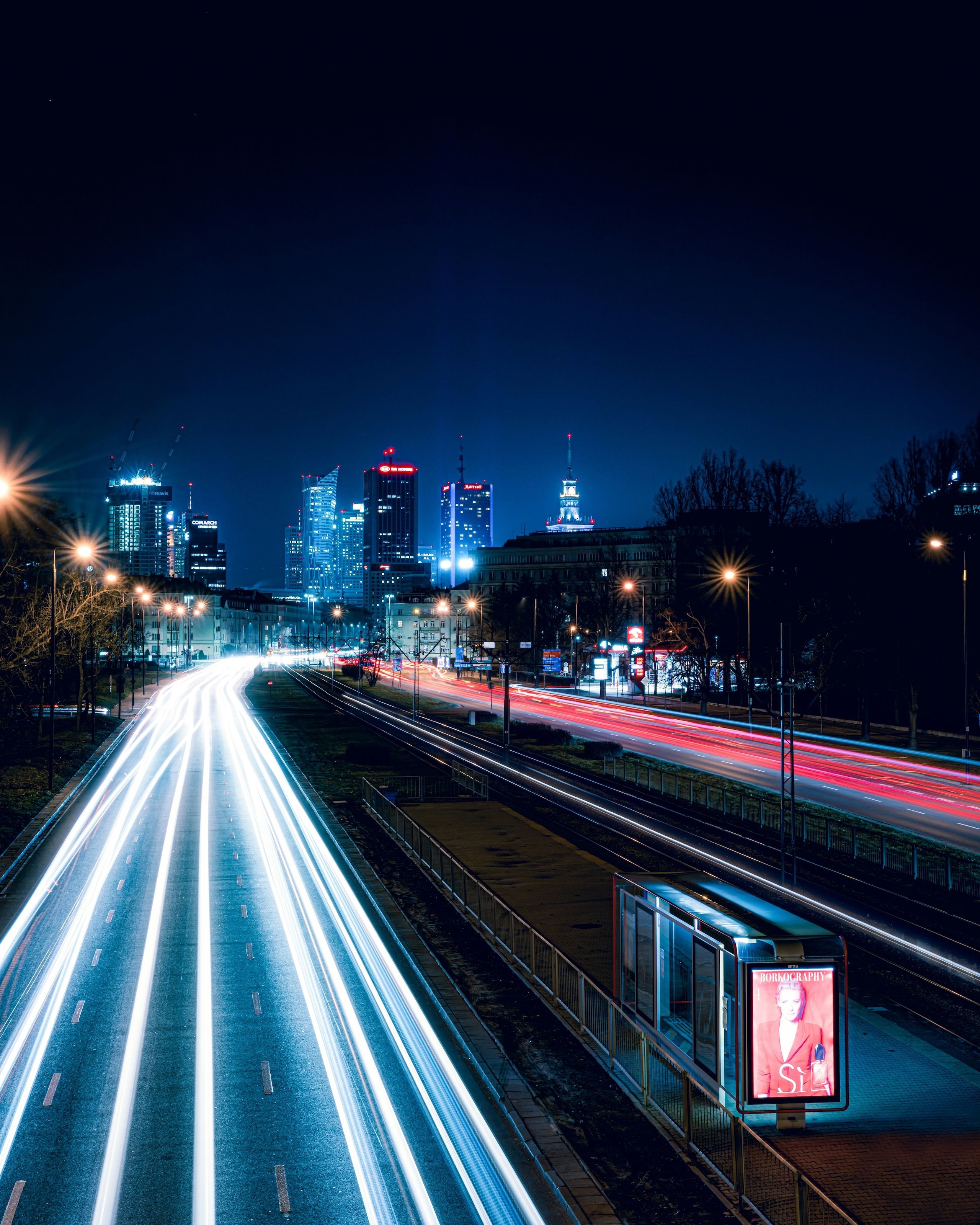 The image shows a busy street with a Warsaw skyline visible in the background. The scene is illuminated by street lights, creating a bustling urban atmosphere at night.