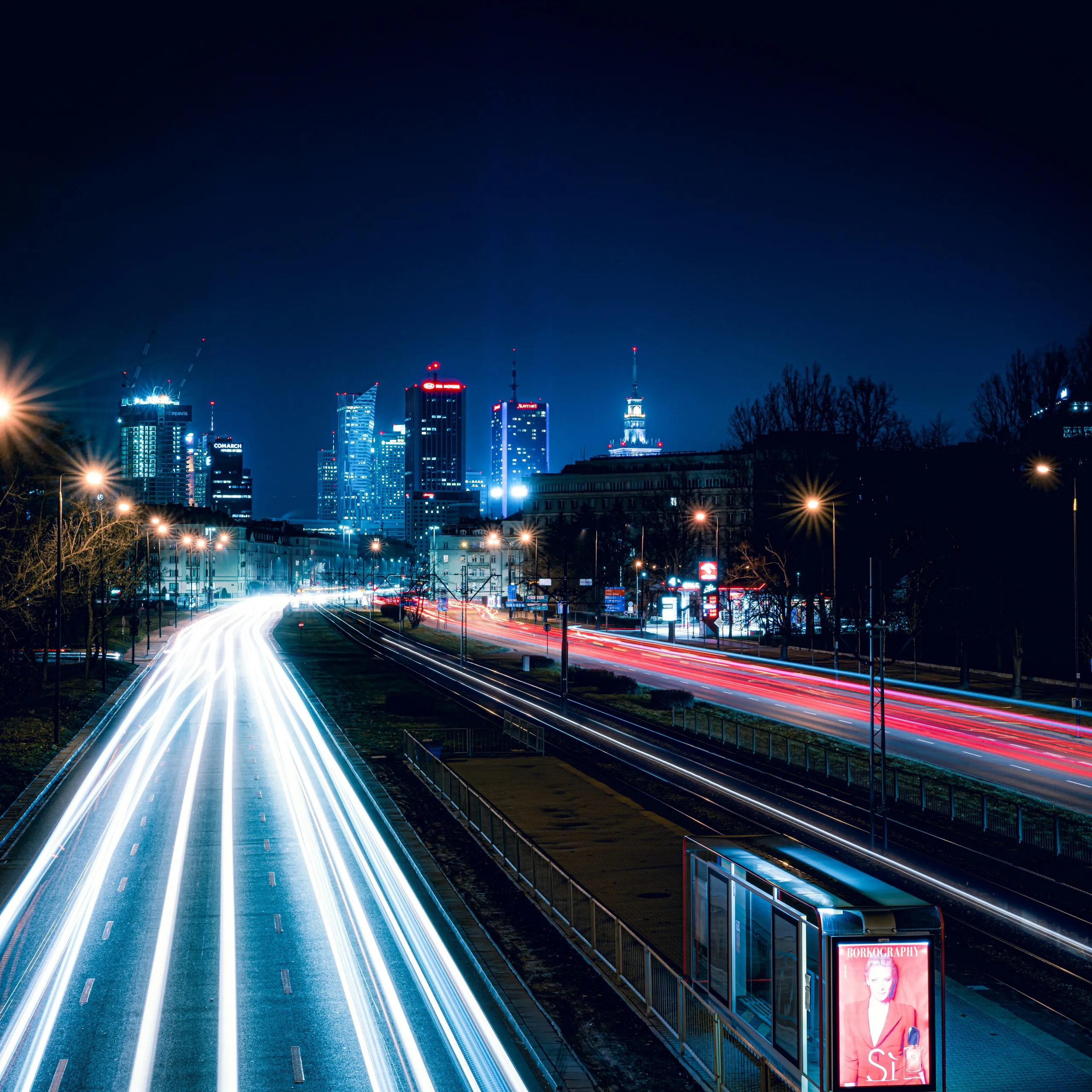 The image shows a highway with a city skyline visible in the background. The scene is illuminated by street lights, creating a bustling urban atmosphere at night.